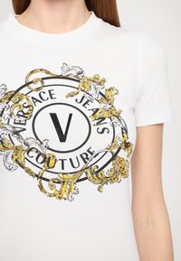 Valge puuvillane T-särk, mille keskosas on ümmargune logo "Versace Jeans Couture", millel on mustad ja kuldsed lillemustrid ning aktsendid.