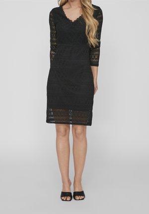 Czarna koronkowa sukienka z długimi rękawami, dekoltem w kształcie litery V oraz dopasowaną sylwetką. Charakteryzuje się skomplikowanymi wzorami i długością do kolan. Czarne buty z odkrytymi palcami.