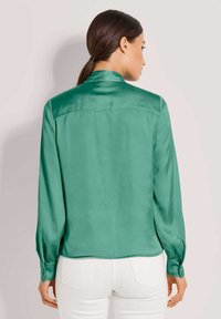 Grüne Satinbluse mit langen Ärmeln, geknöpften Manschetten und einem Rückensattel-Detail. Glatte Textur und leicht abgerundeter Saum.