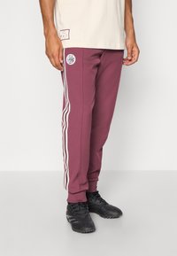 Bordowe spodnie dresowe z białymi paskami po bokach i okrągłym logo na udzie, wykonane z miękkiego materiału o zwężanym kroju. Czarne buty sportowe na nogach.
