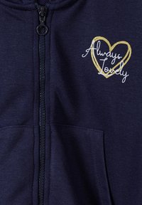 Sudadera con cremallera en azul marino y textura suave, que presenta un gráfico de corazón dorado y un texto en blanco que dice "Siempre Encantadora" en la parte superior izquierda.