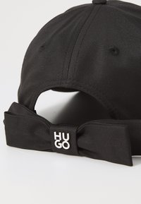 Black cap made of smooth fabric featuring a bow detail at the back, marked with a white "HUGO" logo. Structured shape with ventilation holes.