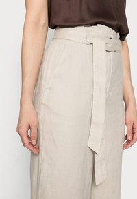 Les pantalons en lin beige clair présentent une taille haute avec une ceinture en tissu, des poches latérales structurées et une coupe ample à jambes larges.