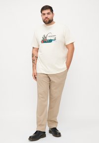 Beige Grafik-T-Shirt mit einem Fischmotiv, kombiniert mit hellen Khaki-Hosen und schwarzen Schnürschuhen, die einen lässigen Look kreieren.