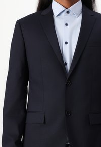 Navy suit jacket with two buttons, notched lapels, and a chest pocket, paired with a light blue button-up shirt featuring dark buttons.