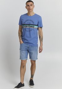 Blå bomull t-skjorte med "California" trykk i svart og turkise striper, parret med lyse denimshorts og svarte sneakers.