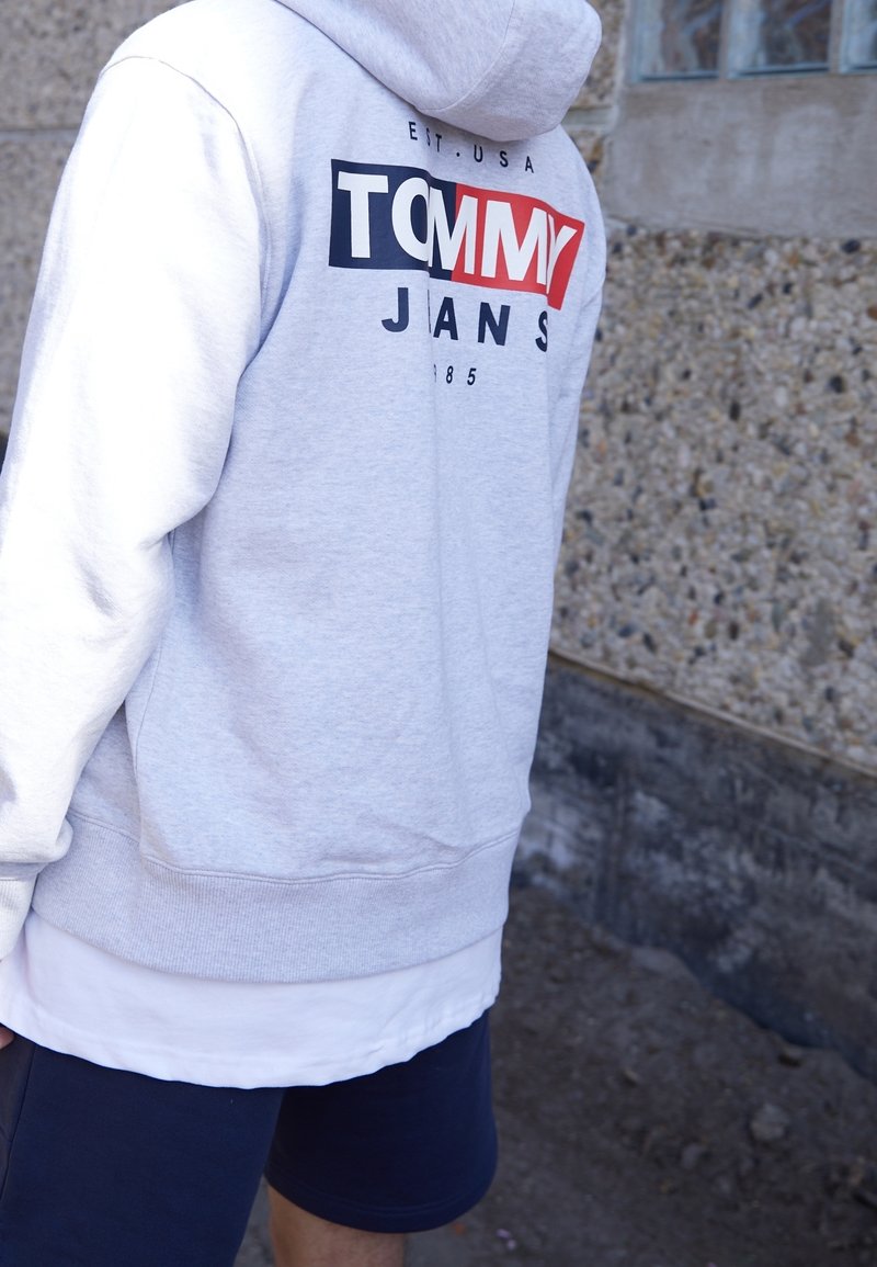 Personne portant un sweat à capuche gris clair avec un logo Tommy Jeans rouge, blanc et bleu, debout à l'extérieur près d'un mur en pierre texturé.