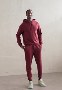 Bărbat îmbrăcat cu un hanorac și pantaloni de trening burgundy, asortați, cu adidași de culoare deschisă, stând împotriva unui perete simplu.