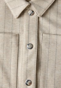 Beige knäppskjorta med en texturerad tyg och tunna vertikala ränder, samt två bröstfickor, säkrade med metallknappar.