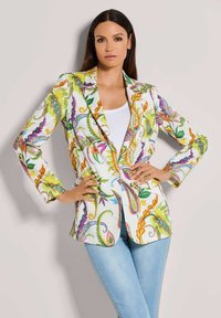 Frau trägt ein farbenfrohes Blazer mit Blumenmuster über einem weißen Oberteil und hellblauen Jeans, posiert mit den Händen auf den Hüften vor einem schlichten Hintergrund.