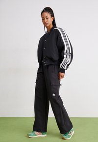Black bomber jacket with white stripes, black cargo pants, and green sneakers with pink accents. Adidas branding on jacket and pants.