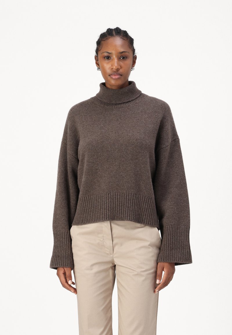 Καφέ χοντρό πλεκτό πουλόβερ με ψηλό λαιμό και κομμένο στυλ, ενώνονται με φαρδιές ριγωτές μανσέτες. Φορεμένο πάνω από ανοιχτό μπεζ παντελόνι, σε λευκό φόντο.