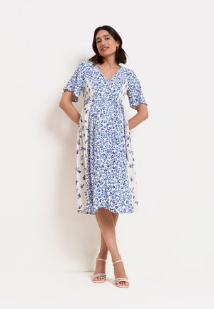Femme portant une robe blanche longueur genou avec un imprimé floral bleu, manches courtes et col en V, debout les mains derrière le dos et portant des sandales blanches.