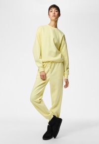 Persoon staand, gekleed in een lichtgele sweatshirt en joggingbroek met zwarte enkellaarzen tegen een effen witte achtergrond.