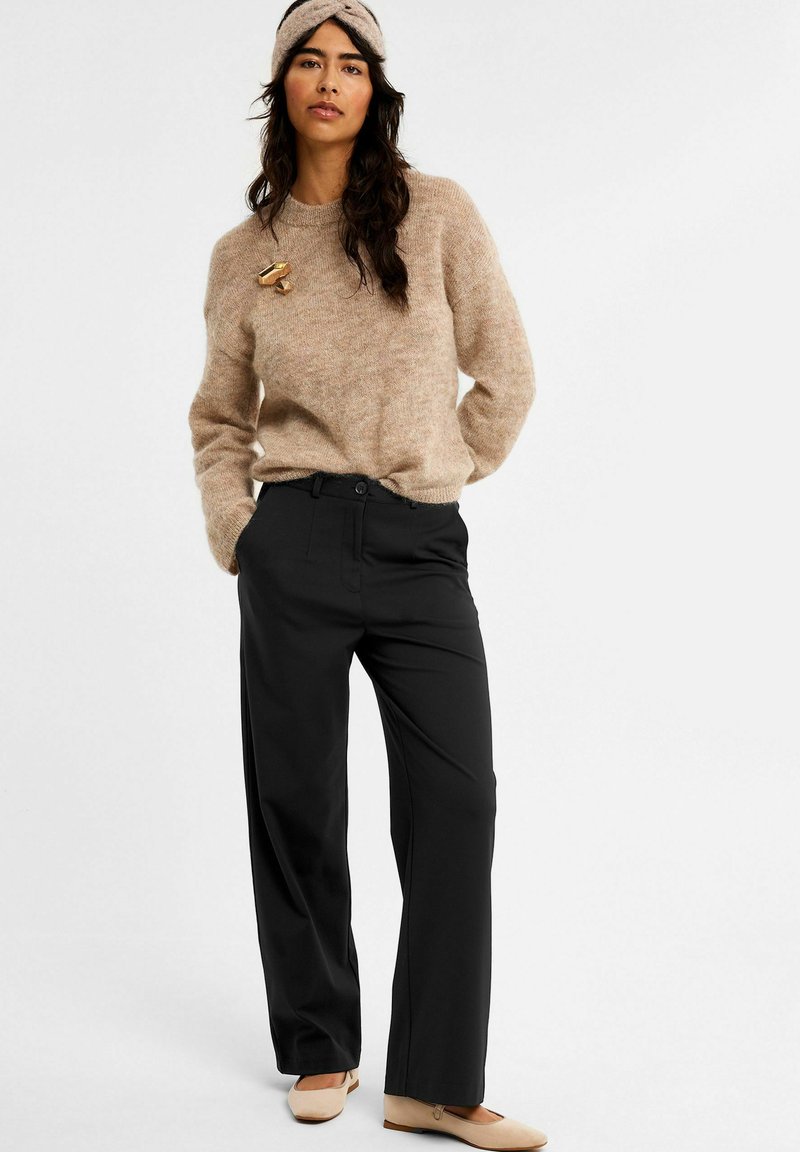 Vrouw in een beige fuzzy trui, zwarte wijde broek, beige ballerina's en een bijpassende beige gebreide haarband, staat met haar handen in haar zakken.