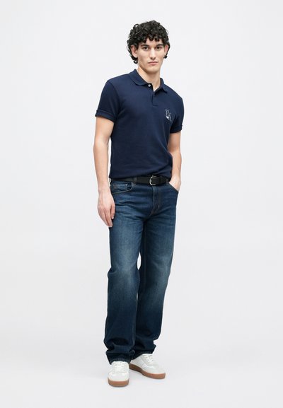 Πόλο μπλούζα σε ναυτικό μπλε με κοντά μανίκια και διακριτική επωνυμία, συνδυασμένη με τζιν μεσαίου μπλε χρώματος. Το μοντέλο φοράει λευκά αθλητικά παπούτσια και μαύρη ζώνη.