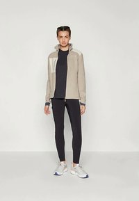 Young person standing against plain background wearing beige zip-up jacket, black leggings, black shirt, and white sneakers with orange accents.