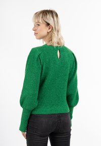 Maglione verde a maglia con tessuto testurizzato, maniche a palloncino e dettaglio a chiave sulla schiena fissato da un bottone verde. Indossato con jeans neri.