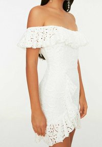 Valkoinen olkaimeton mekko pitsidetaleilla, röyhelöillä varustetuilla hihoilla, istuvalla bodysuitilla ja aaltoilevalla helman reunalla. Teksturoitu kukkakuosi koko mekossa.