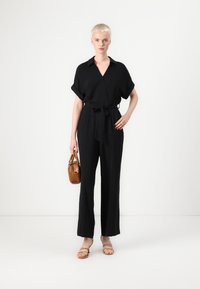 Svart jumpsuit med dyp V-hals, korte oppbrettede ermer og knyttet liv. Kombinert med en brun håndveske og sandal med stropper.