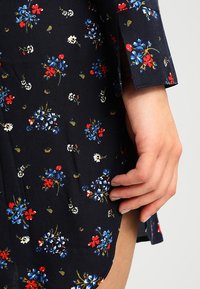 Květinové šaty z tmavě modré látky s drobnými červenými, modrými a bílými květy. Dlouhé rukávy s rozparkem a volný střih.