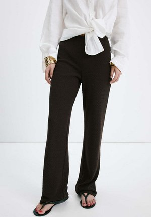 Osoba nosiaca čierne nohavice so širokými nohavicami, bielu previazanú blúzku, zlaté náramky a čierne žabky s červeným lakom na nechtoch na nohách, stojaca na bielej podlahe.