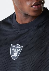 Maillot de sport en mailles noires avec un col rond, des trous de ventilation et un logo des Raiders blanc à l'avant. Tissu texturé avec un design élégant.