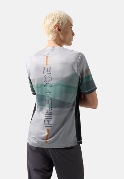 Junge Person mit kurzen blonden Haaren, die ein grau-grünes gemustertes T-Shirt mit dem Text „FIRE+ICE“ auf dem Rücken trägt und mit verschränkten Armen steht.