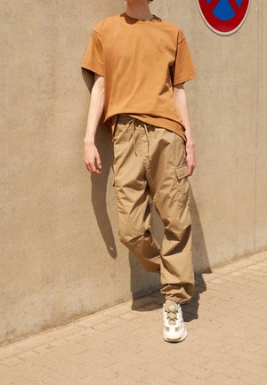 Person iført brown t-shirt, beige cargo bukser og hvide sneakers, der går på en belagt fortov ved siden af en betonvæg med et parkeringsforbudsskilt.