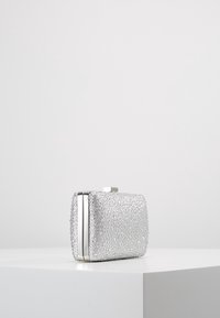 Petit sac à main rectangulaire argenté recouvert de strass scintillants, avec un cadre et un fermoir en métal, sur un piédestal blanc face à un fond clair.