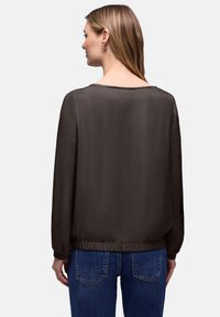 Blouse en satin marron foncé à manches longues, ourlet élastique froncé et large décolleté, associée à un jean bleu. Texture lisse avec un léger éclat subtil.