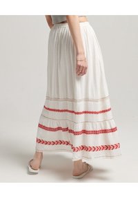 Witte maxi-rok met rode geborduurde patronen en kanten rand, voorzien van een geplooide tailleband en lichtgewicht stof. Platte sandalen zichtbaar.