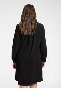 Abito nero a maniche lunghe con colletto, caratterizzato da una linea dritta, tessuto morbido e polsini con bottoni. Vista posteriore con capelli che scendono lungo la schiena.