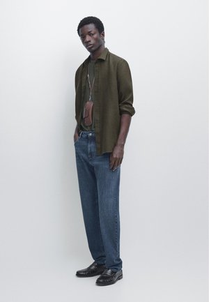 Man wearing olive green shirt over green tee, blue jeans, black loafers, and brown leather pouch hanging from neck, standing against white background.
