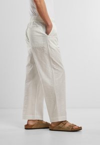 Svetlé tkané nohavice s textúrovaným vertikálnym vzorom, s bočnými vreckami a voľným strihom, doladené s hnedými sandálmi.