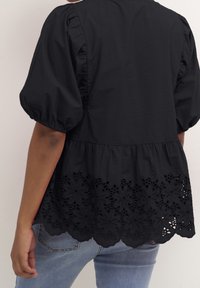 Musta paita puffeilla hihoilla ja scalloped-helmalla, jossa on monimutkaista kukkakirjontaa alaosassa. Sujuva kangas, rento istuvuus.