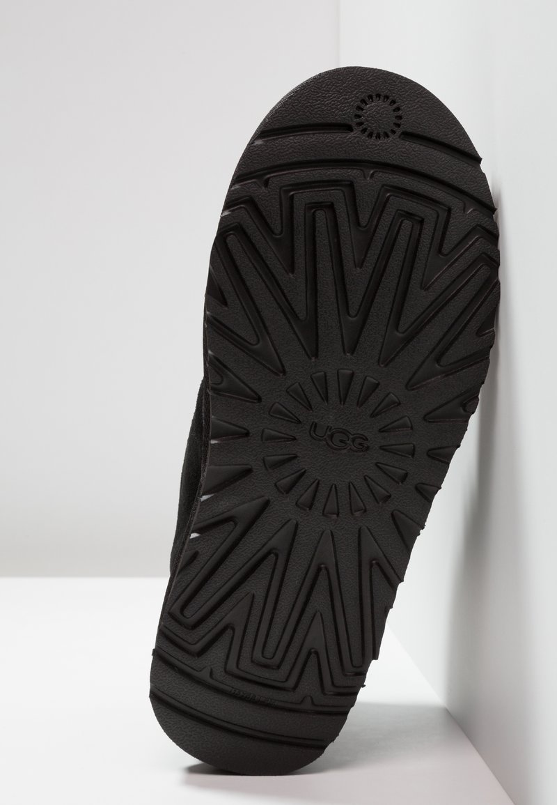 Black rubber sole with a textured pattern, featuring angular grooves and a circular logo at the center, positioned against a light background.