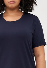 Curly-haired person wearing a navy blue t-shirt with a rounded neckline, focusing on the upper chest and face. Simple, casual attire.