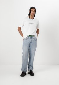 T-shirt bianca in cotone con logo stampato, abbinata a jeans larghi blu chiaro e scarpe platform nere. Modello in posa in piedi con le mani in tasca.