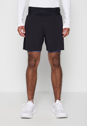 Homme portant un short de sport noir superposé, un haut à manches longues blanc et des chaussures de course blanches, debout devant un fond uni.