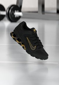 Sapatilha de treino Nike em preto com detalhes em dourado. Apresenta um upper texturizado, design de amarração e uma sola acolchoada com unidades de ar visíveis.