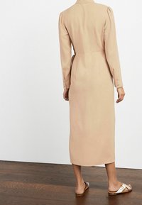 Donna che indossa un lungo vestito beige con bottoni sui polsini, abbinato a sandali bianchi, su un pavimento in legno, di spalle.
