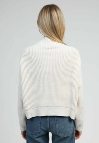 Pull cropped en tricot blanc avec une texture côtelée, présentant un col montant et des épaules tombantes, associé à un jean bleu taille haute.