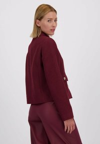 Vrouw met blond haar in een getextureerde bordeauxrode blazer en bijpassende broek, die over haar schouder kijkt tegen een effen witte achtergrond.