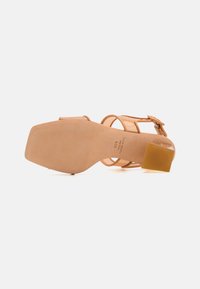Beige high-heeled sandal with transparent straps, square molded heel, and smooth suede texture. Features a buckle closure on the ankle.