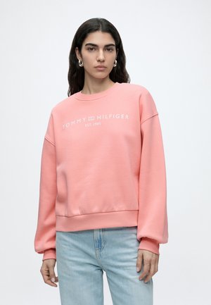 Kobieta o ciemnych włosach nosi różową bluzę Tommy Hilfiger i jasnoniebieskie dżinsy, stoi na jednolitym jasnym tle.