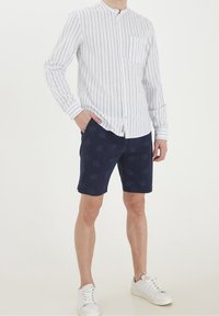 Camisa blanca de manga larga con rayas verticales azules, combinada con pantalones cortos navy con un patrón de palmera. Zapatillas blancas completan el atuendo.