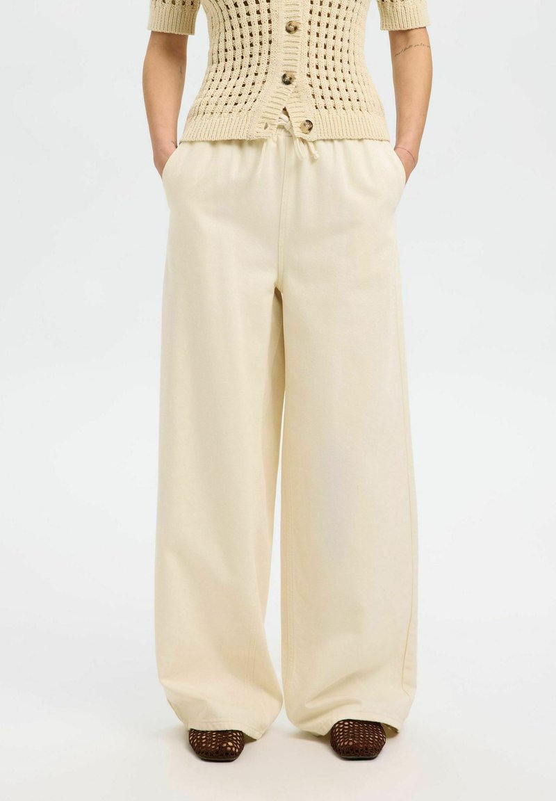 Pantalons larges couleur crème avec une taille élastique, confectionnés en tissu doux. Présente des poches latérales et une coupe décontractée pour le confort.