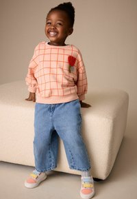 Meruňková mikina se vzorem mřížky a červenou výšivkou květin, ve spojení s volnými modrými džínami a vícebarevnými teniskami, stojící na světlém povrchu.