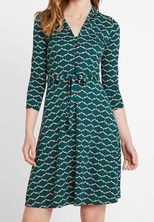 Teal dress with geometric chain-link pattern, three-quarter sleeves, buttoned front, and waist tie belt, worn by a standing person.